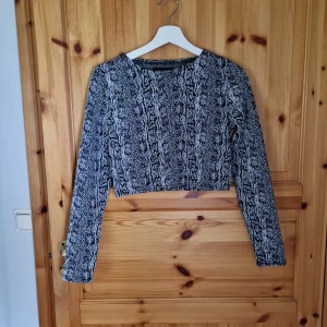 Svartvit mönstrad croppad tröja - Säljer en svartvit croppad tröja med ett unikt mönster som påminner om ormskinn. Tröjan har långa ärmar och är perfekt för en trendig look. Passar bra till både vardag och fest!