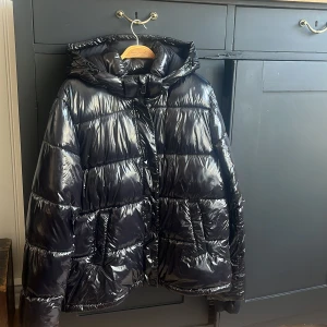 Svart glansig pufferjacka från ONLY - Säljer en supercool svart glansig pufferjacka från ONLY i storlek M. Jackan har en quiltad design med dragkedja och knappar framtill. Perfekt för att hålla sig varm och stilren under kalla dagar. Den har en huva för extra skydd mot väder och vind. Passar perfekt för både vardag och festliga tillfällen!