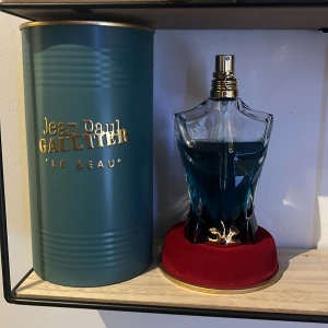 Jean Paul Gaultier Le Beau Parfym - Säljer en supercool parfym från Jean Paul Gaultier som heter 'Le Beau'. Flaskan är formad som en manlig torso i en snygg blå färg och kommer med en lyxig röd bas. Förpackningen är en stilren grön cylinder med guldtext. Perfekt för den som vill ha en unik och elegant doftupplevelse!