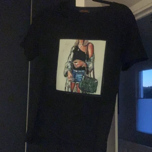 Svart t-shirt med tryck från Orange - Säljer en svart t-shirt från märket Orange i storlek S. Den har ett coolt tryck på framsidan med en stilfull illustration av en person i trendiga kläder. Perfekt för en casual look eller att styla upp med en jacka. T-shirten är i bra skick och redo för nya äventyr!