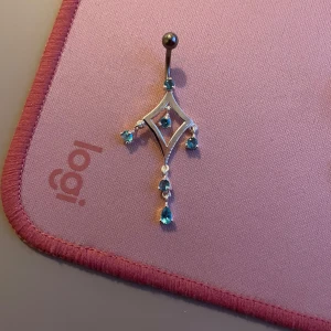 Snygg navelpiercing med blå stenar - Elegant navelpiercing i silver med en unik diamantformad design. Den är dekorerad med flera små blå stenar som hänger i kedjor, vilket ger ett stilrent och glittrigt utseende. Perfekt för att lägga till en touch av glamour.
