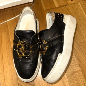 Svarta sneakers från Roberto Cavalli - Svarta sneakers i skinn från Roberto Cavalli med gulddetaljer och nitar. Skorna har en unik design med ormskinnsmönster på hälen och en dekorativ guldfärgad spänne framtill. Sällsynta tillverkas ej mer!