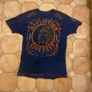 Blå t-shirt från Affliction - Säljer en blå t-shirt från Affliction med tryck av en indianhuvudbonad och texten 'Live Fast'. T-shirten har korta ärmar och orangea detaljer. Perfekt för en avslappnad stil.