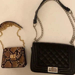 Svart quiltad axelväska och ormmönstrad handväska - Säljer en stilren svart quiltad axelväska med silverkedja och en trendig ormmönstrad handväska med guldkedja. Perfekta accessoarer för att lyfta din outfit. Den lilla kostar 40kr och den svarta 70kr! 
