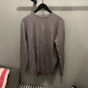 Grå tröja från Lyle & Scott - Snygg grå tröja från Lyle & Scott med deras ikoniska logga på bröstet. Perfekt för en stilren och avslappnad look. Tröjan har långa ärmar och en rund halsringning.