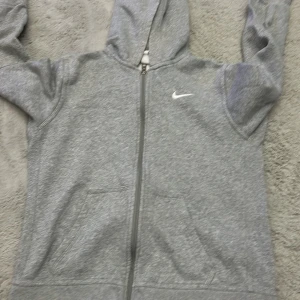 Grå hoodie från Nike - Säljer en grå hoodie från Nike med dragkedja och huva. Den har en klassisk design med fickor framtill och det ikoniska Nike-loggan på bröstet. Perfekt för en avslappnad stil.