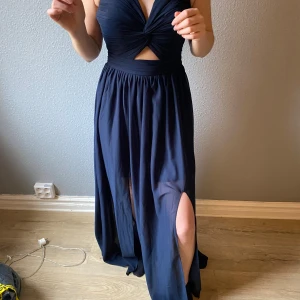 Mörkblå långklänning med knyt - Elegant mörkblå långklänning med djup v-ringning och knyt i ryggen. Klänningen har en slits framtill och är ärmlös, vilket ger en stilren look. Perfekt för speciella tillfällen. Perfekt till balen.