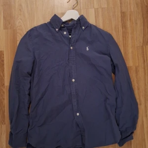 Blå skjorta från Ralph Lauren - Snygg blå långärmad skjorta från Ralph Lauren med broderad logga på bröstet. Skjortan har en avslappnad passform och knappar framtill. Perfekt för en stilren look.