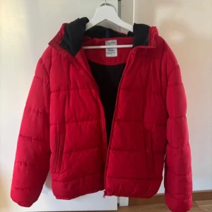 Röd pufferjacka från Divided - Snygg röd pufferjacka från H&M med huva och dragkedja. Perfekt för kyliga dagar. Jackan har en stilren look och är lätt att matcha med olika outfits. Köpt för flera år sedan men inte använd så mycket :) Som man kan se på sista bilden har den två pytte små hål, vill du ha bättre bilder så skriv :)