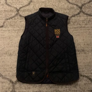 Mörkblå quiltad väst från Morris - Snygg mörkblå quiltad väst från Morris med broderad logotyp och brittisk flagga på bröstet. Västen har en dragkedja framtill och är perfekt för lager-på-lager under kyligare dagar.