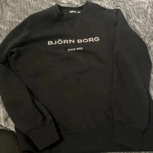 Svart tröja från Björn Borg - Säljer en svart tröja från Björn Borg med texten 'BJÖRN BORG SINCE 1956' på framsidan. Tröjan har långa ärmar och en klassisk rund halsringning. Perfekt för en avslappnad stil. Den är också använd men i bra skick fortfarande!