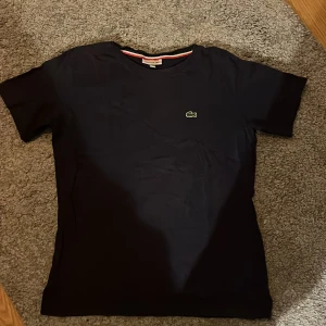 Mörkblå t-shirt från Lacoste - Säljer en klassisk mörkblå t-shirt från Lacoste med det ikoniska krokodilemblemet på bröstet. T-shirten har en rund halsringning med en röd och vit randig detalj på insidan av kragen. Perfekt för en stilren och avslappnad look.