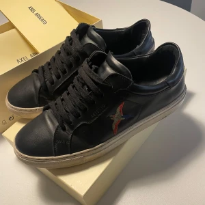 Svarta sneakers från Axel Arigato - Snygga svarta sneakers i skinn från Axel Arigato med broderad fågel på sidan. Skorna har klassisk snörning och en vit sula som ger en stilren look. Perfekta för en trendig vardagsstil. Nypris 2700kr