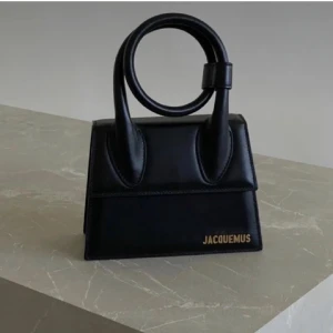 Svart handväska från Jacquemus - Snygg och stilren svart handväska från Jacquemus i skinn. Väskan har ett unikt handtag och guldlogga framtill. Perfekt för att ge din outfit en lyxig touch. Tillkommer en dustbag
