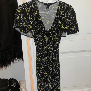 Svart kortklänning med citronmönster - Trendig svart kortklänning med ett lekfullt citronmönster. Klänningen har en v-ringad hals och korta ärmar, perfekt för sommarens varma dagar. Den har en figursydd passform som framhäver midjan.