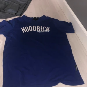 Blå t-shirt från Hoodrich - Säljer en blå t-shirt från Hoodrich med tryck på både fram- och baksidan. T-shirten har korta ärmar och är i storlek XL för åldern 13-15 år. Perfekt för en avslappnad stil. Passar också som S