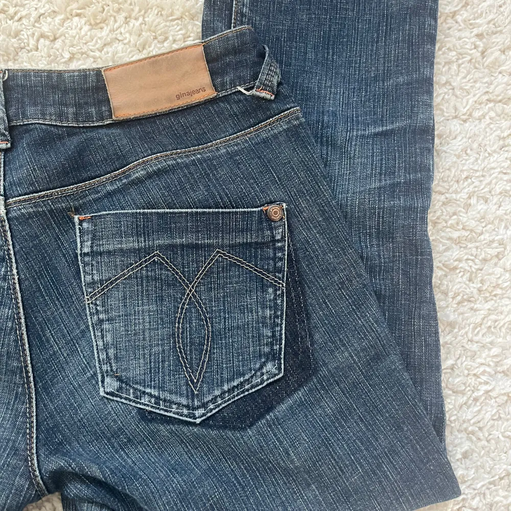 Säljer ett par mörkblå jeans från Gina Jeans med bootcut-stil. De har en klassisk femficksdesign och en snygg detalj på bakfickorna. Jeansen är lågmidjade och har en rå kant vid bensluten för en trendig look.. Farkut & Housut.