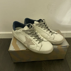 Golden Goose sneakers - Säljer ett par vita Golden Goose Super-Star sneakers med grå snörning och en stjärndetalj på sidan. Skorna har en mörkblå häl och är tillverkade i skinn. Dem är i skick 7/10 då dem är slitna vid hälarna. Storlek 41(passar även42). Dustbag medföljer! Hör av er vid funderingar och frågor🙌