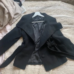 Svart kappa från H&M - Snygg svart kappa från H&M med bred krage och knytbälte i midjan. Perfekt för kyligare dagar med sin stilrena design och bekväma passform.