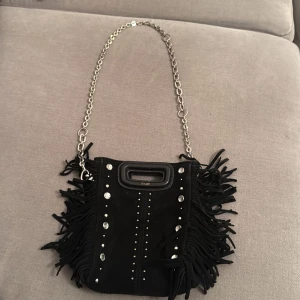 Svart axelväska med fransar och kedja - Jättesnygg Maje väska den är svart i mocka med fransar och silverdetaljer. Väskan har en kedja som axelrem och dekorativa nitar framtill. Perfekt för att ge din outfit en edgy touch. Det är den lilla modellen 