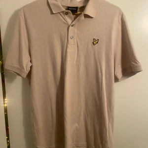 Beige pikétröja från Lyle & Scott - Säljer en stilren beige pikétröja från Lyle & Scott med korta ärmar och klassisk krage. Tröjan har en broderad gul logotyp på bröstet och knappar framtill. Perfekt för en avslappnad men snygg look.