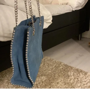 Blå axelväska med kedjedetaljer - Snygg blå axelväska med silverfärgade kedjor som axelremmar och dekorativa nitar längs kanterna. Perfekt för att ge din outfit en cool touch.
