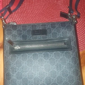 Axelväska från Gucci - Snygg axelväska från Gucci i mörkblått med mönster och en praktisk dragkedja framtill. Väskan har en justerbar axelrem i rött och blått, vilket ger en stilren look. Perfekt för att bära med sig det nödvändigaste med stil.