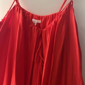 Röd klänning med knytband - Snygg röd klänning med smala axelband och knytband framtill. Den har en luftig passform och volangdetaljer nedtill som ger en fin rörelse. Perfekt för varma sommardagar. Aldrig använd 