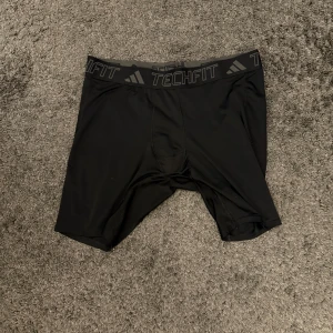 Svarta träningsshorts från Adidas - Säljer ett par svarta träningsshorts från Adidas Techfit. De är tillverkade i ett stretchigt och bekvämt material, perfekt för intensiva träningspass. Shortsen har en elastisk midja med Techfit-logga. Använda oftast som underställ, storlek XL