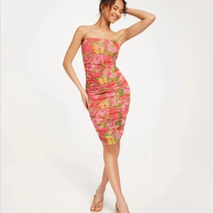 Färgglad axelbandslös klänning - Snygg axelbandslös klänning med ett färgglatt mönster i rosa, grönt och gult. Klänningen har en tight passform och är perfekt för sommarens festligheter. Den är tillverkad i ett mjukt och stretchigt material som ger en bekväm känsla. Aldrig använd. Köpt på Nelly 