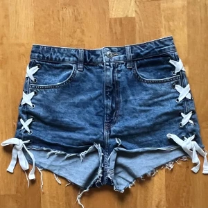 Blå jeansshorts med snörning - Snygga blå jeansshorts med cool snörning på sidorna och råa kanter. Perfekta för en avslappnad stil med en twist. De har en klassisk femficksdesign och knappgylf.