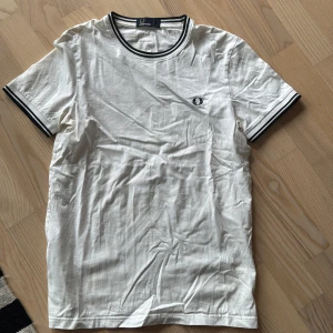 Vit t-shirt från Fred Perry - Säljer en klassisk vit t-shirt från Fred Perry med svarta och vita ränder på krage och ärmslut. T-shirten har en broderad logga på bröstet och är tillverkad i bomull. Perfekt för en stilren look!