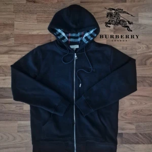 Burberry kofta - Snygg svart hoodie från Burberry med dragkedja och klassiskt rutigt mönster på insidan av huvan. Den har långa ärmar och en broderad logga på bröstet. Perfekt för en stilren look.