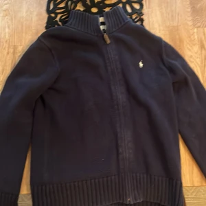 Mörkblå kofta från Ralph Lauren - Snygg mörkblå kofta från Ralph Lauren med dragkedja och ribbade detaljer. Perfekt för kyligare dagar och ger en stilren look. Den har en liten broderad logga på bröstet.Storlek 14-16 år