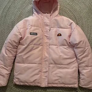Rosa pufferjacka från Ellesse - Snygg rosa pufferjacka från Ellesse med huva och dragkedja. Jackan är tillverkad i 100% polyester och har en broderad logga på bröstet. Perfekt för kyliga dagar.  Oanvänd som ny. Original pris ca 1000kr