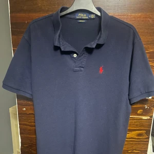 Mörkblå pikétröja från Ralph Lauren - Säljer en klassisk mörkblå pikétröja från Ralph Lauren med röd logga på bröstet. Tröjan har korta ärmar och en stilren krage. Perfekt för en avslappnad men stilfull look. Skick 9/10