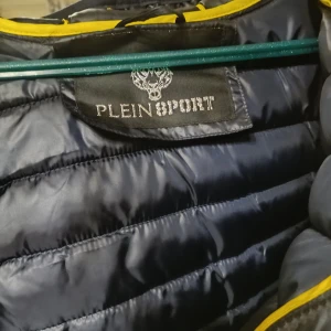 Svart pufferjacka från Plein Sport - Snygg svart pufferjacka från Plein Sport med gul logga på ryggen och huvan. Jackan har dragkedja framtill och två sidofickor med dragkedjor. Perfekt för kyliga dagar med sin quiltade design.