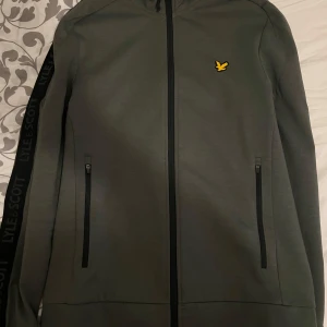 MörkGrön kofta från Lyle & Scott sport - Snygg grön kofta från Lyle & Scott med dragkedja och långärmad design. Koftan har ett diskret märke på bröstet och svarta detaljer längs ärmarna. Perfekt för en stilren look.