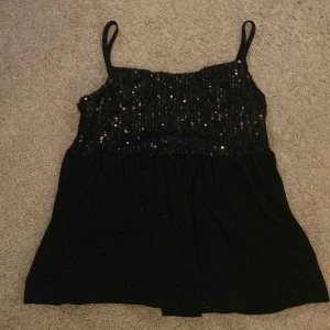 Svart paljettlinne från Gina Tricot - Snyggt svart linne från Gina Tricot med glittriga paljetter på överdelen. Linnet har smala axelband och en luftig passform. Säljer för att jag har växt ur den.