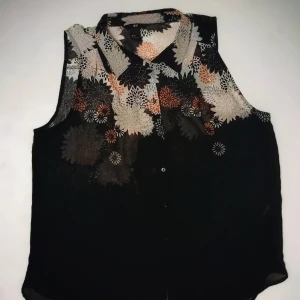 Blommig ärmlös blus - Snygg ärmlös blus med blommönster i svart, beige och orange. Perfekt för en stilren look med krage och knappar framtill.