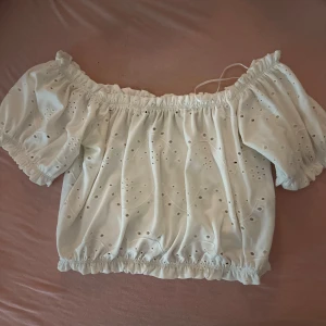 Vit offshoulder topp från Gina Tricot - Säljer en söt vit offshoulder topp från Gina Tricot. Toppen har ett broderat mönster och elastiska kanter vid ärmar och midja för en bekväm passform. Perfekt för varma sommardagar!