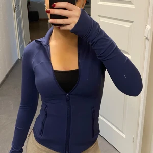 Mörkblå träningskofta med dragkedja - Snygg mörkblå träningskofta med dragkedja och hål till tummen. Finns även en ficka vid armen där man kan ha airpods eller nycklar medans man tränar. Sitter som en smäck och bra material. Oanvänd och prislapp kvar