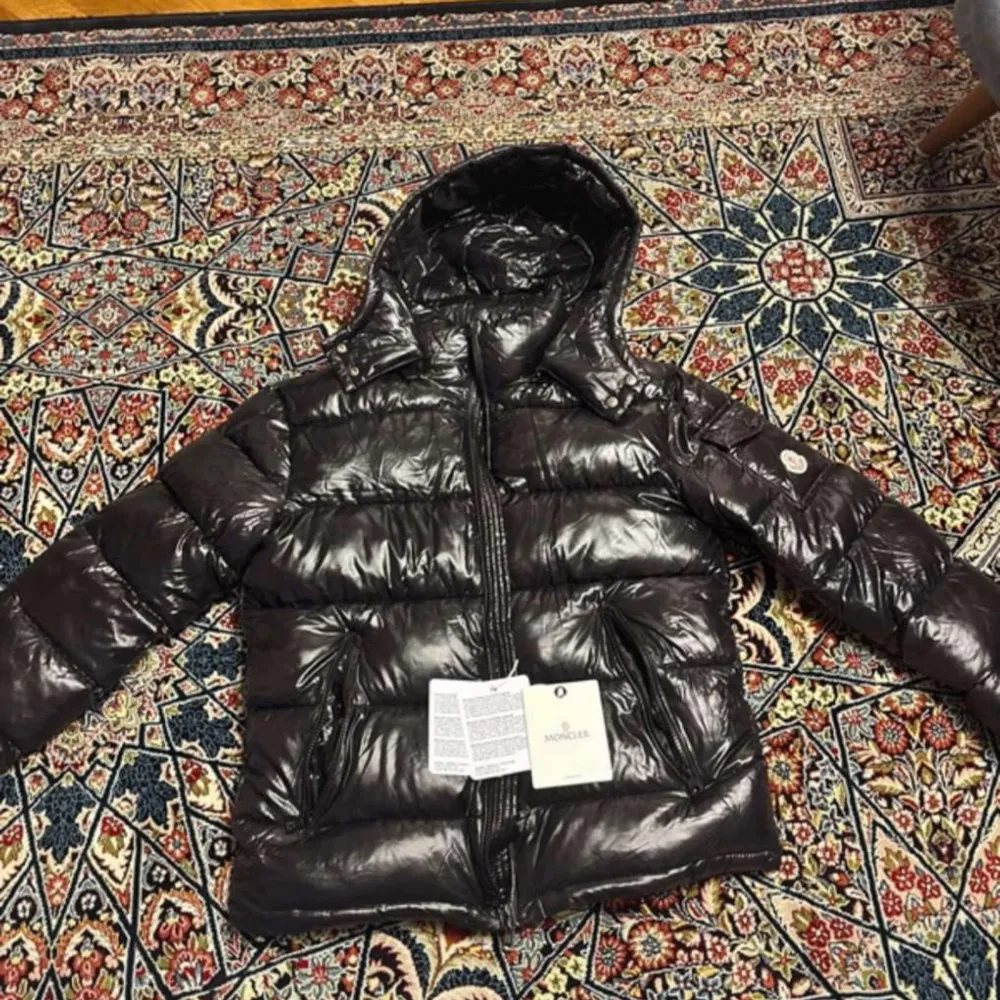 Snygg och varm svart dunjacka från Moncler med glansig finish. Jackan har en praktisk huva och dragkedja framtill. Perfekt för kyliga dagar.. Takit.