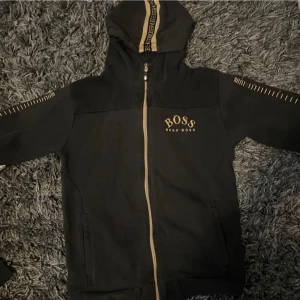 Svart hoodie från Hugo Boss - Snygg svart hoodie från Hugo Boss med dragkedja och huva. Den har guldfärgade detaljer och logotyp på bröstet. Perfekt för en stilren och bekväm look.