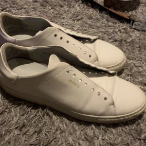 Vita sneakers från Axel Arigato - Säljer ett par stilrena vita sneakers från Axel Arigato. Skorna är tillverkade i skinn och har en minimalistisk design utan snörning. Perfekta för en avslappnad och trendig look.