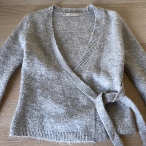 Grå omlottkofta från Gina Tricot - Varan är aldrig använd. Säljer en mysig grå omlottkofta från Gina Tricot i premiumkvalitet. Koftan har långa ärmar och ett knytband i midjan för en snygg passform. Perfekt för kyliga dagar!