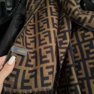Brun och svart halsduk från Fendi - Snygg och elegant halsduk från Fendi i brunt och svart med det ikoniska Fendi-mönstret. Perfekt accessoar för att lyfta din outfit med en touch av lyx. Den är lång och har fransar i ändarna.