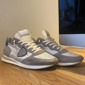 Grå och vita sneakers från Philippe Model - Snygga sneakers från Philippe Model i grått och vitt med blå detaljer. Skorna har en klassisk design med snörning och en bekväm sula. Perfekta för en stilren look.