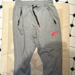 Grå mjukisbyxor  - Säljer ett par grå mjukisbyxor med Nike i storlek S. De har en bekväm passform med snörning i midjan och ett rött Nike-logotyptryck på benet. Perfekta för träning eller en avslappnad dag hemma. 