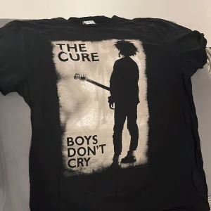 Svart T-shirt med The Cure-tryck - Cool svart T-shirt från Amplified med tryck av The Cure och texten 'Boys Don't Cry'. Perfekt för fans av bandet eller för en rockig stil. T-shirten har en normal passform och är kortärmad, vilket gör den idealisk för vardagsbruk.